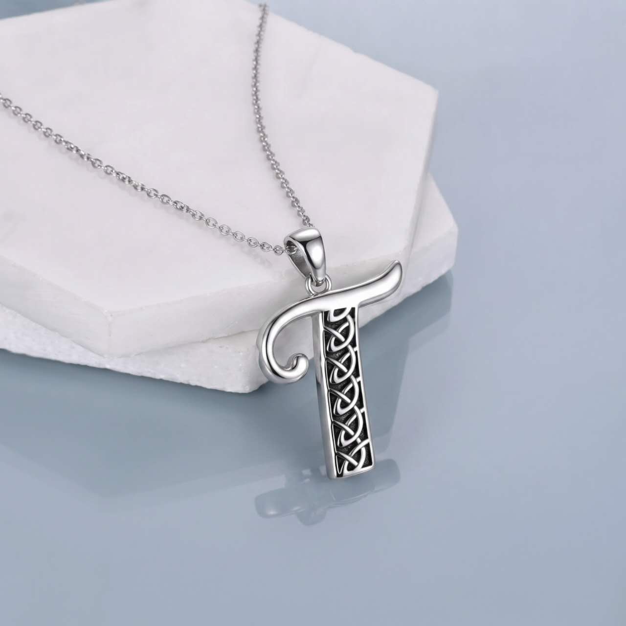 A silver pendant necklace featuring the letter 'T' with an intricate Celtic knot design inside, resting on a white textured display block against a light blue-gray reflective background.