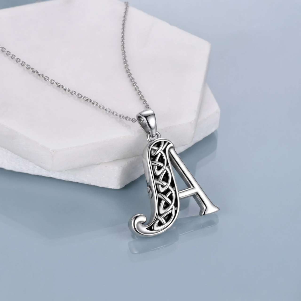 A silver Celtic-style letter 'A' pendant necklace resting on a white geometric display block against a light blue-gray background.