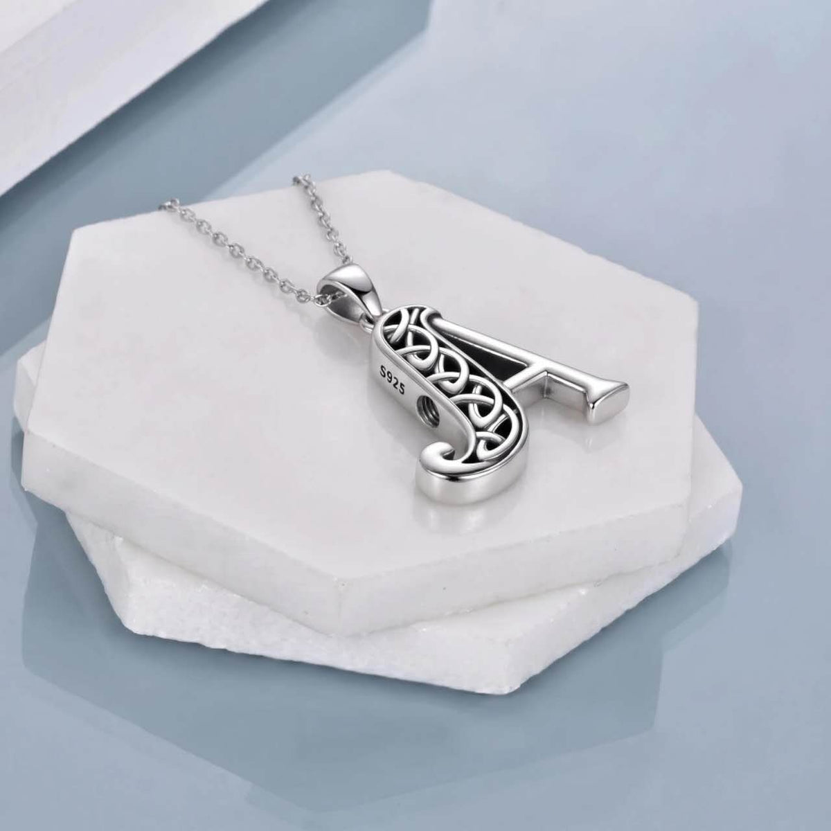 A silver initial pendant necklace spelling 'A' with an intricate Celtic knot design, resting on a white hexagonal stone display against a light blue-gray background.