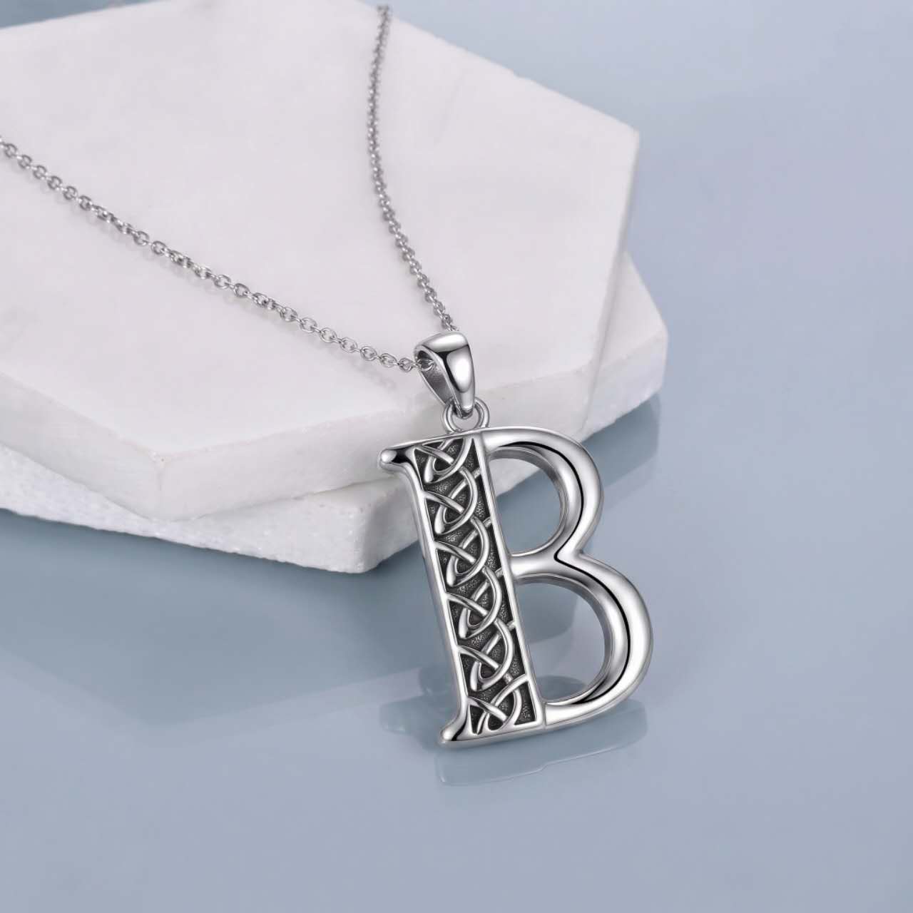 A silver initial pendant necklace featuring the letter 'B' with an intricate Celtic knot design on the vertical stem, resting on a white hexagonal display block against a light blue-gray background.