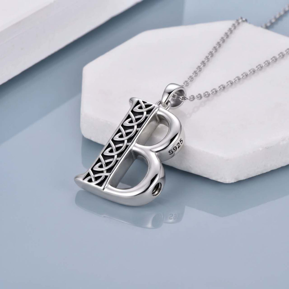 A sterling silver initial pendant necklace featuring the letter 'B' with an intricate Celtic knot design on one side and a smooth polished silver on the other, resting on a white hexagonal display block.