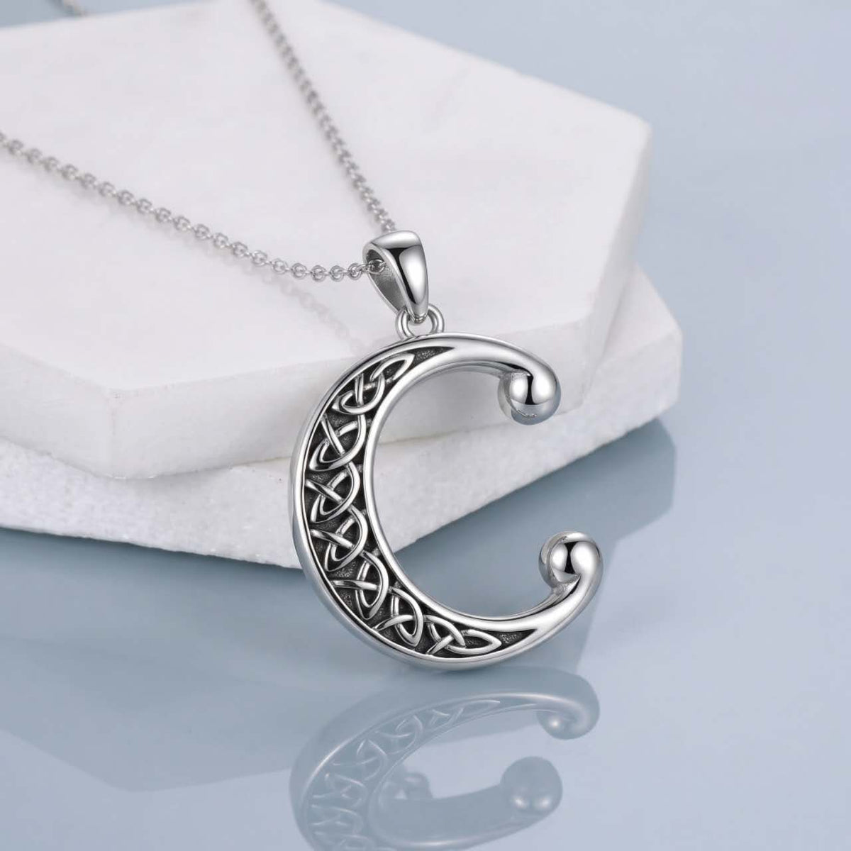 A silver crescent moon pendant necklace with an intricate Celtic knot pattern, resting on a white display block over a light blue reflective surface.