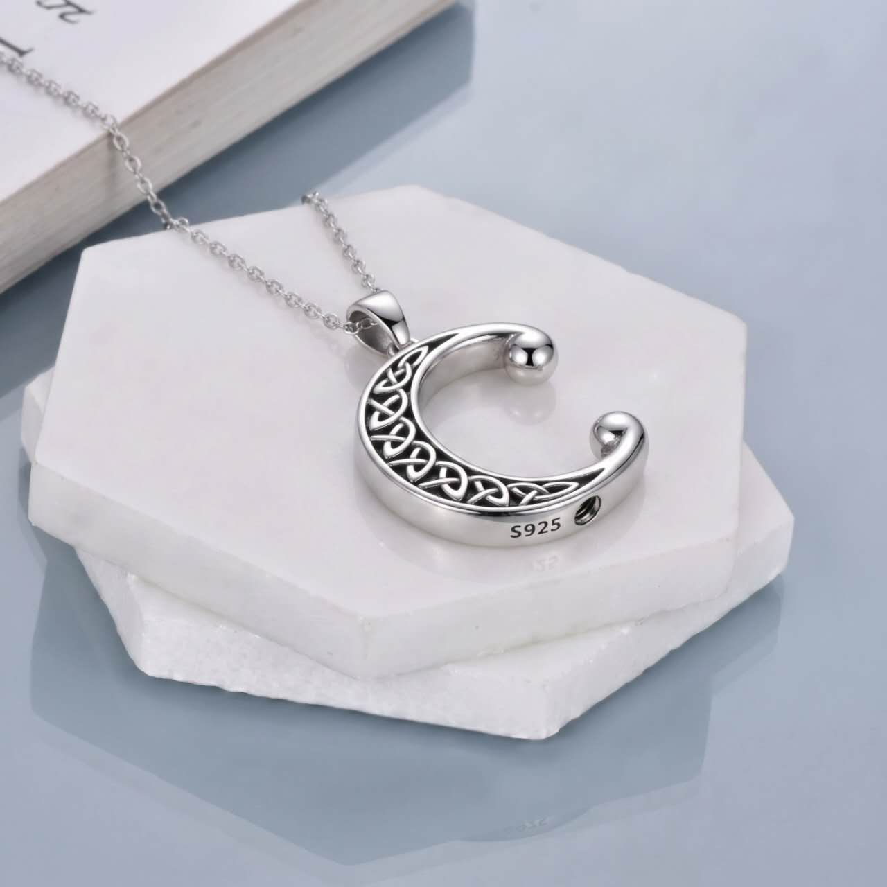 A silver crescent moon pendant necklace with an intricate filigree cutout pattern and a polished ball at each end, resting on stacked white hexagonal stone slabs. The necklace is marked 'S925' and features a delicate silver chain.