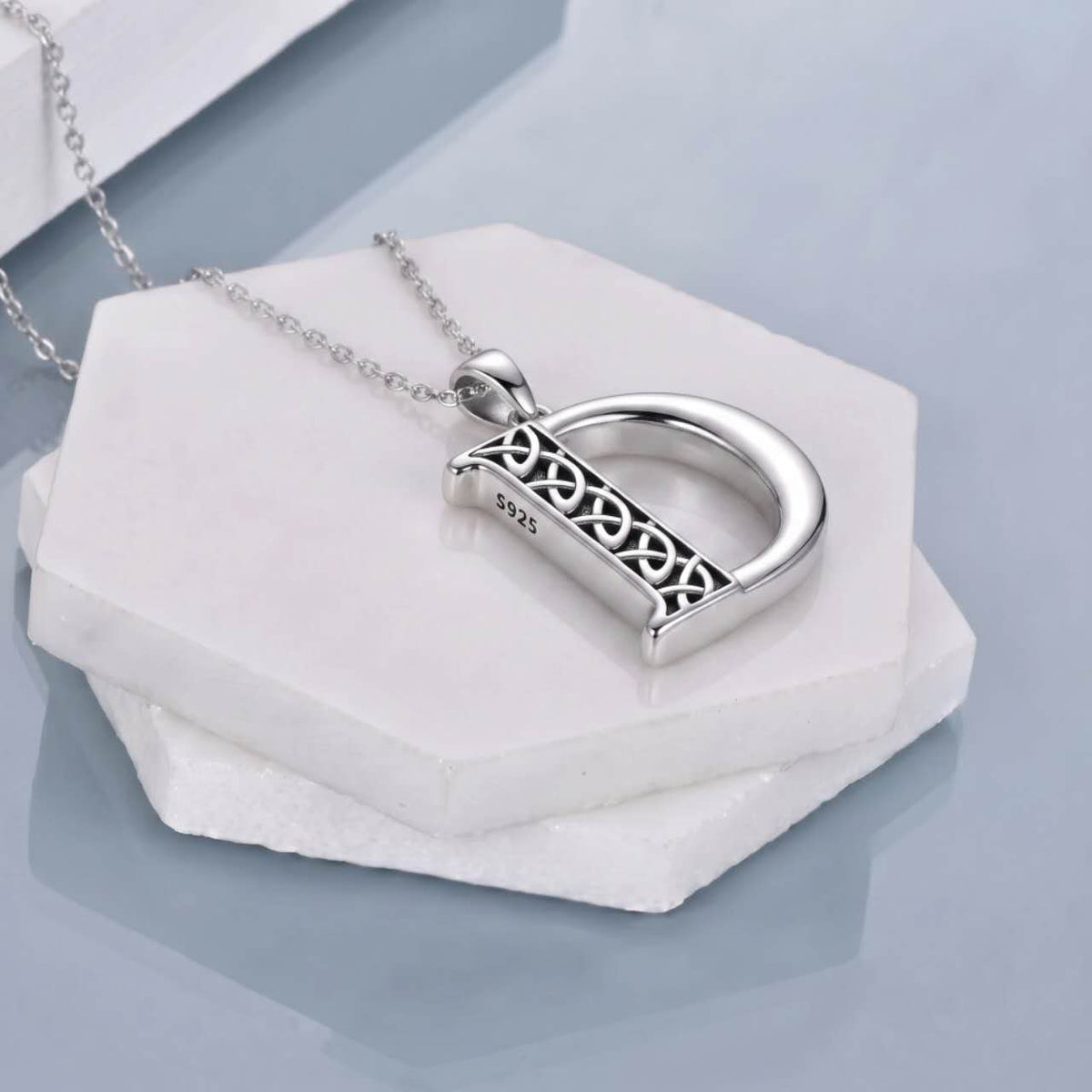 A silver initial pendant necklace spelling 'D' with an Celtic knot design side, resting on stacked white hexagonal stone blocks against a cool light blue-gray background.