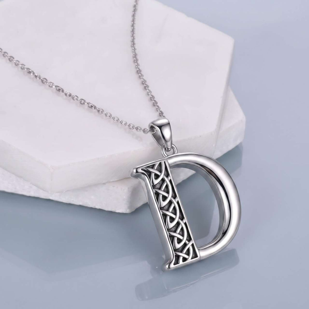 A silver initial pendant necklace featuring the letter 'D' with an intricate Celtic knot design on its left side, resting on a white geometric display block against a light blue-gray background.