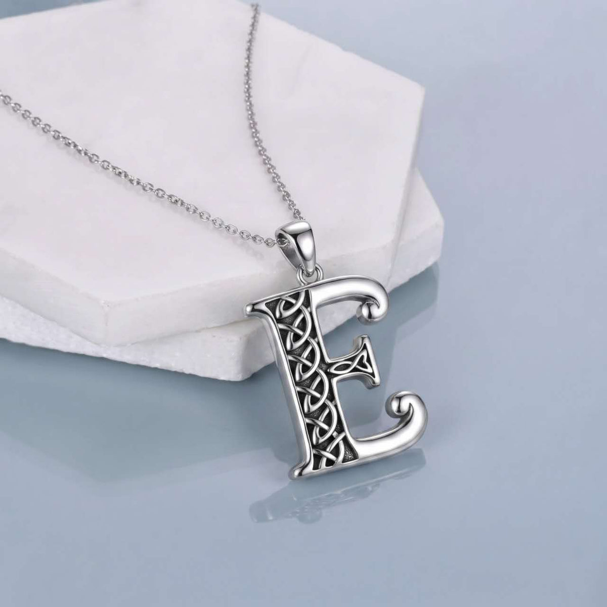 A silver Celtic-style letter 'E' pendant necklace resting on a white geometric display block against a light blue-gray background.