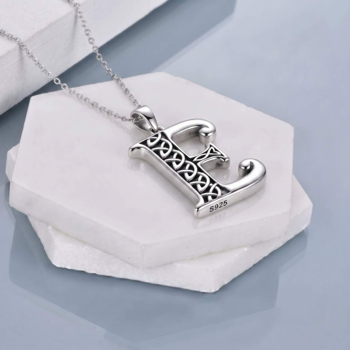 A sterling silver Celtic letter 'E' pendant necklace on a delicate silver chain, displayed on stacked white hexagonal stone slabs.