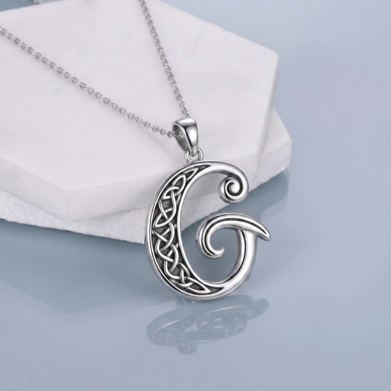 A silver Celtic letter 'G' pendant necklace with an intricate knotwork design, resting on a white display block against a light blue-gray reflective background.