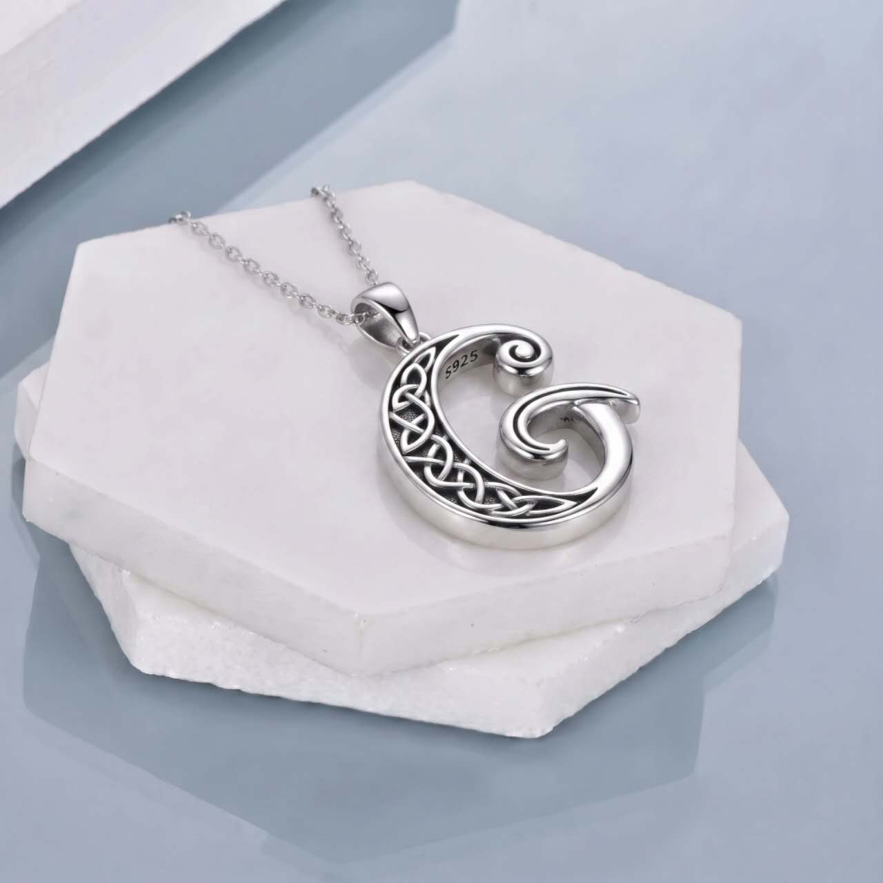 A silver pendant necklace with a Celtic knot design and a spiral motif, resting on a white hexagonal stone block against a light blue-gray background.