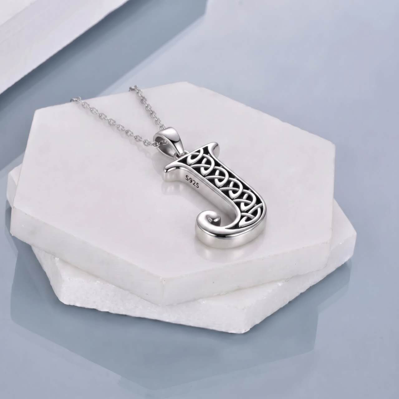A silver Celtic knot cremation pendant necklace featuring the letter 'J' on a delicate silver chain, displayed on stacked white stone slabs against a cool gray background.