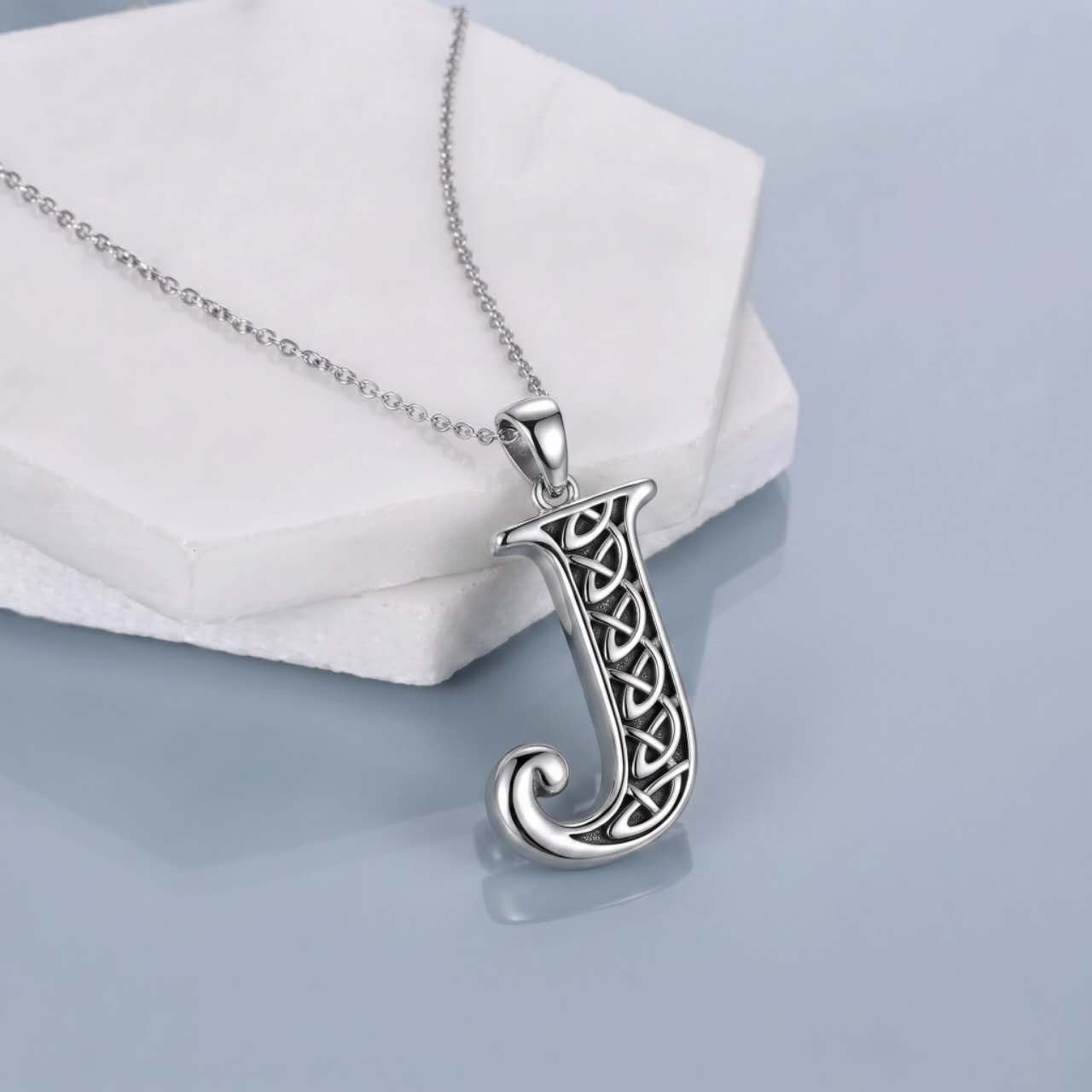 A silver initial pendant necklace featuring the letter 'J' with an intricate Celtic knot design, resting on a white geometric display block against a light blue-gray background.