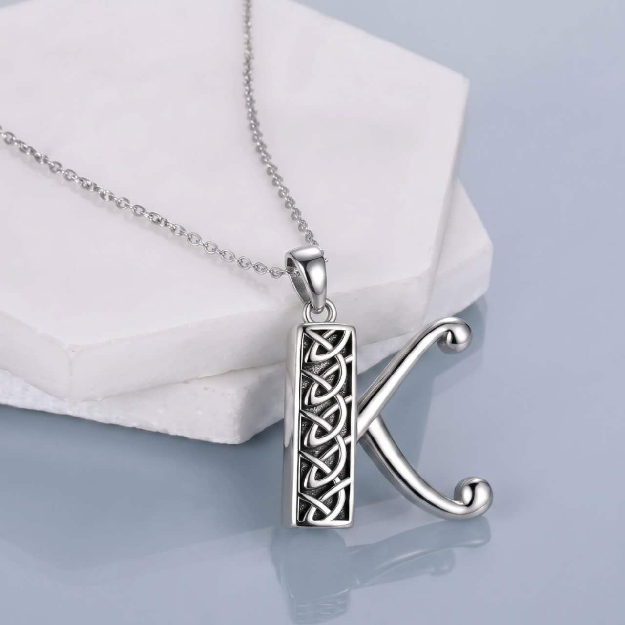 A silver initial pendant necklace featuring the letter 'K' with an integrated Celtic knot design, resting on a white geometric display block against a light blue-gray background.