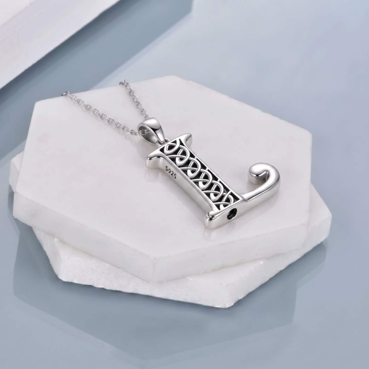 A silver-toned pendant necklace with a stylized letter 'L' featuring an intricate Celtic knot pattern, resting on a white hexagonal stone display against a light blue-gray background.