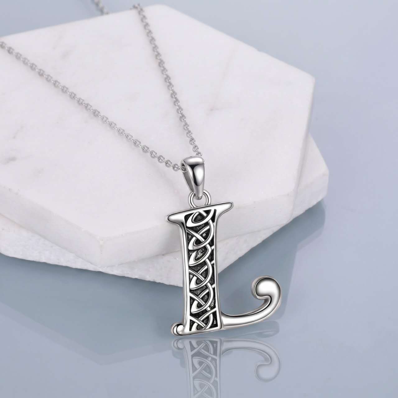 A silver Celtic-style letter 'L' pendant necklace on a delicate silver chain, displayed on a white marble hexagonal block against a light gray reflective background.