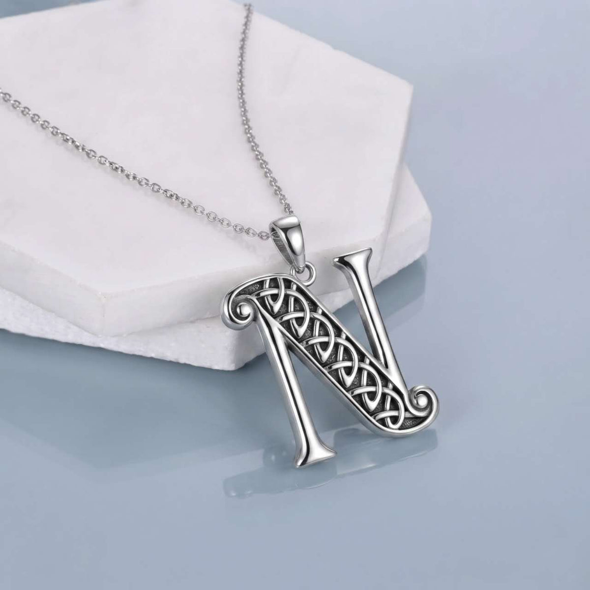 A silver initial pendant necklace featuring the letter 'N' with an intricate Celtic knot design, resting on a white textured display block against a light blue-gray background.