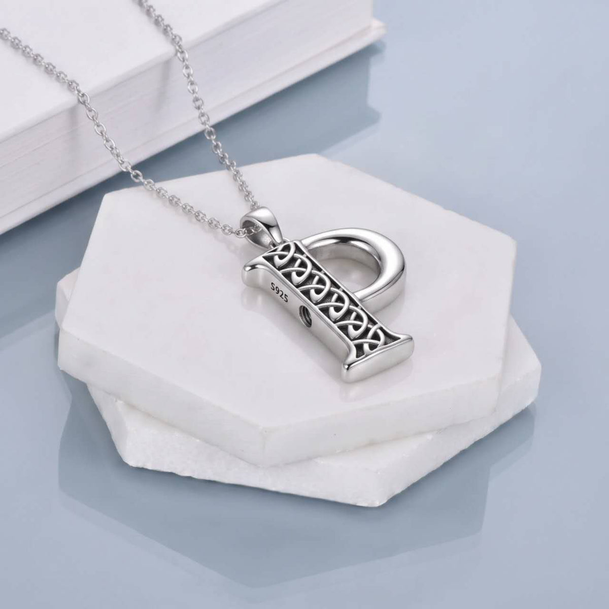 A silver pendant P necklace featuring an ornate Celtic knotwork design on a rectangular bar, attached to a delicate silver chain. The necklace is displayed on a white hexagonal stone stand with a white book in the background, set against a light blue-gray reflective surface.