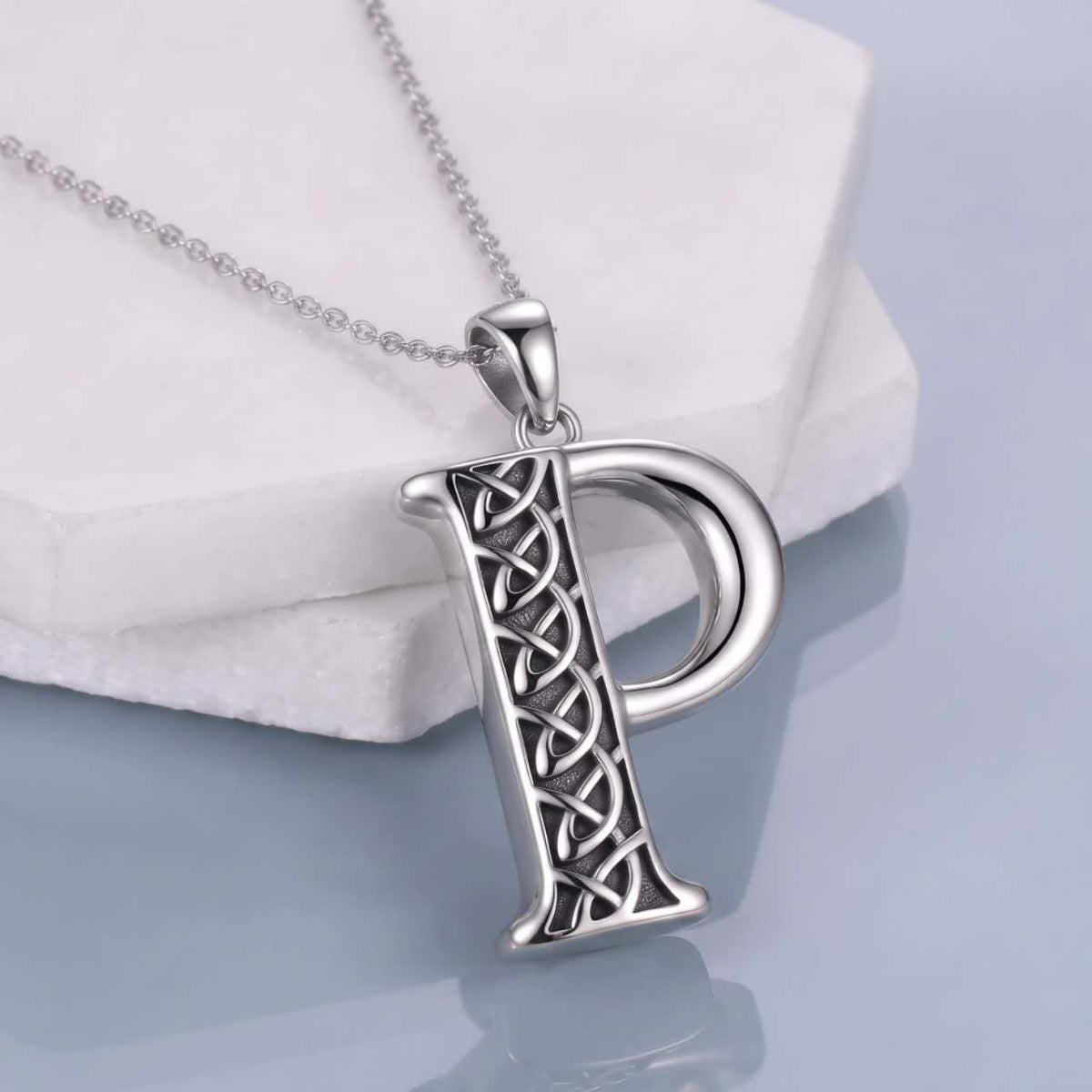 A silver initial pendant necklace featuring the letter 'P' with an intricate Celtic knot design on its stem, suspended from a delicate silver chain and displayed on a white stone block.