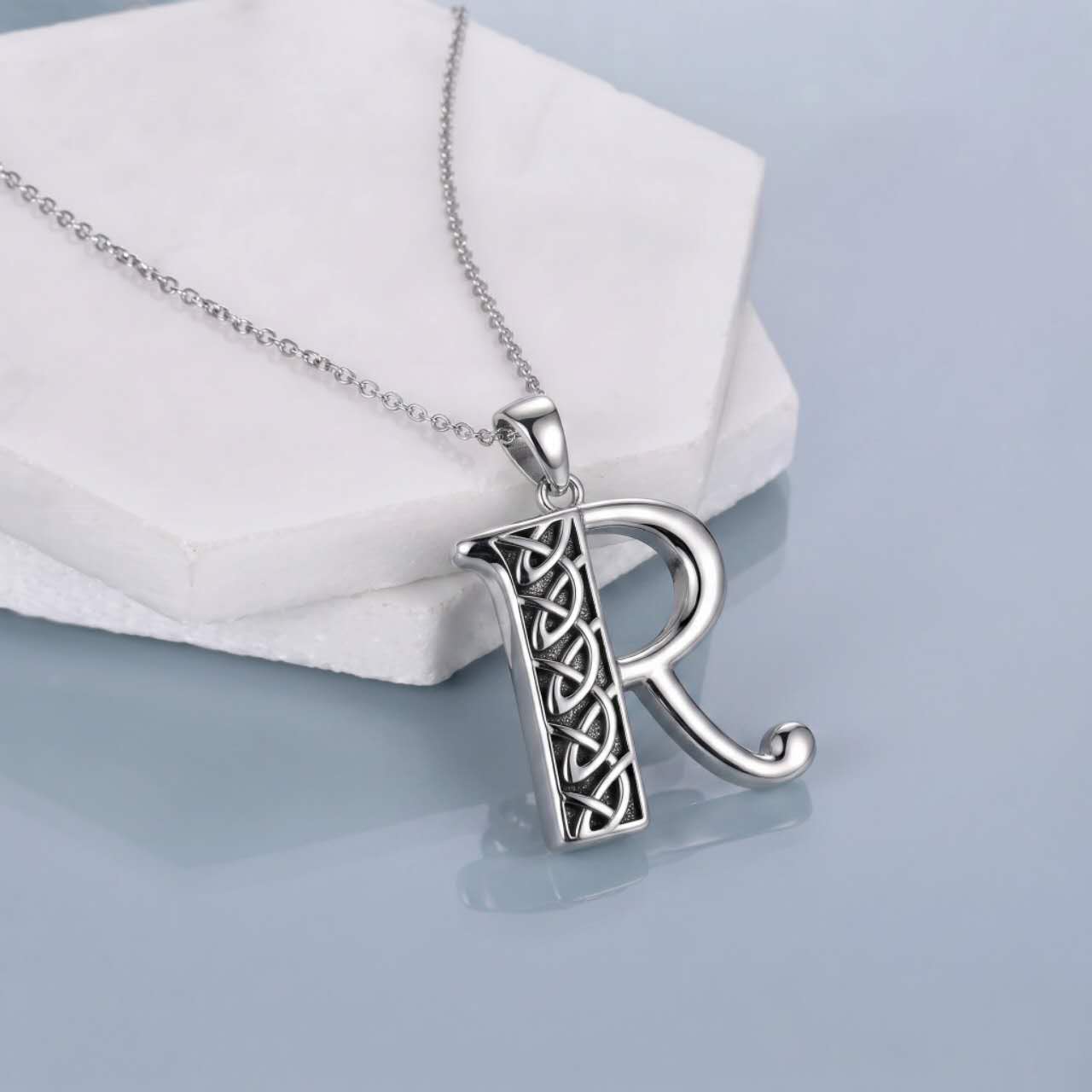 A sterling silver initial pendant necklace featuring the letter 'R' with a Celtic knot design, resting on a white geometric display block against a light blue-gray background.