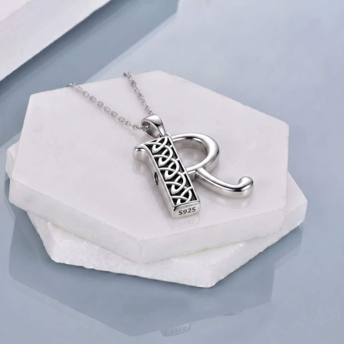 A sterling silver letter R cremation necklace featuring a Celtic knot design, displayed on a white hexagonal stone block against a light gray background.