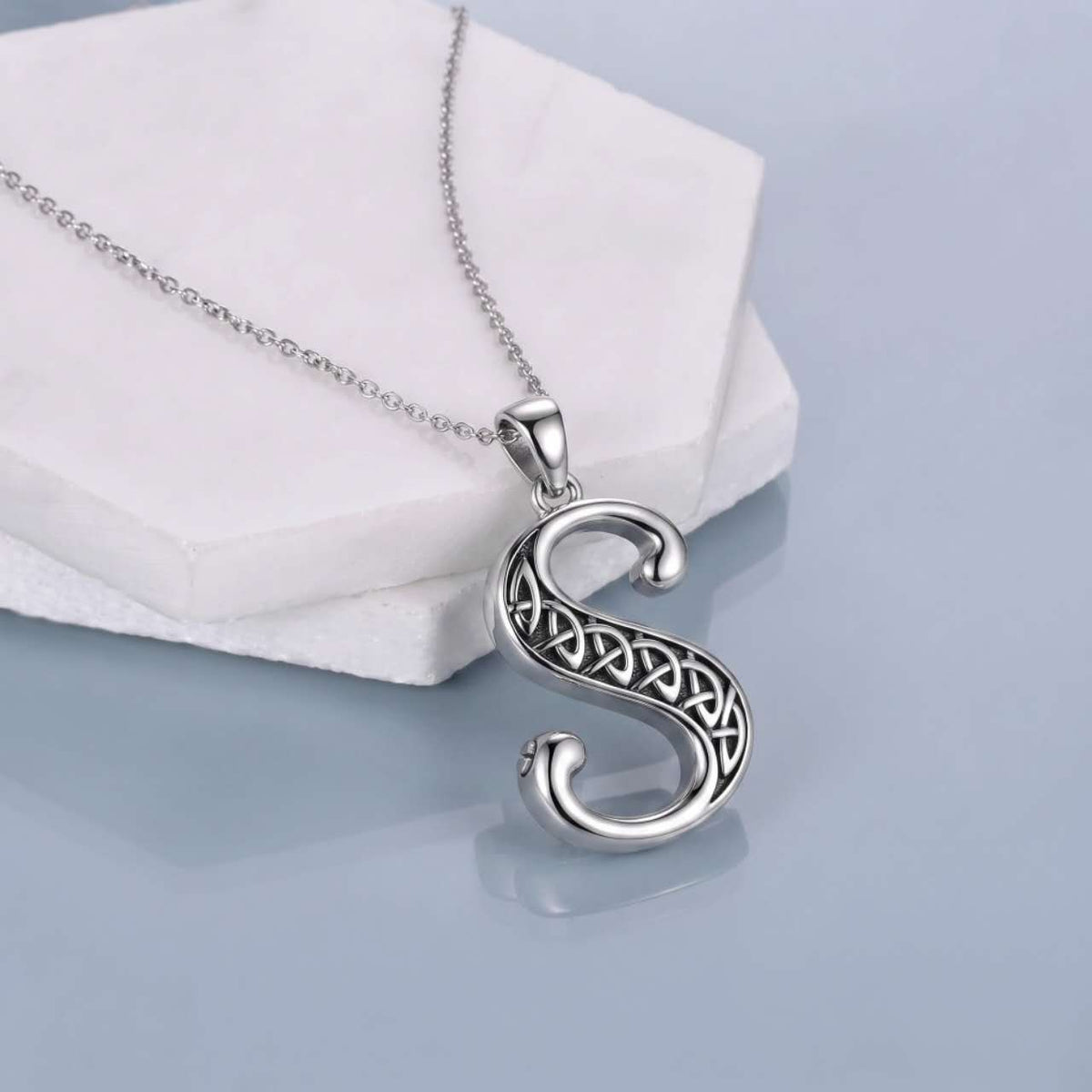 A silver Celtic-style letter 'S' pendant necklace with an engraved knot pattern, resting on a white geometric display block against a light blue-gray background.