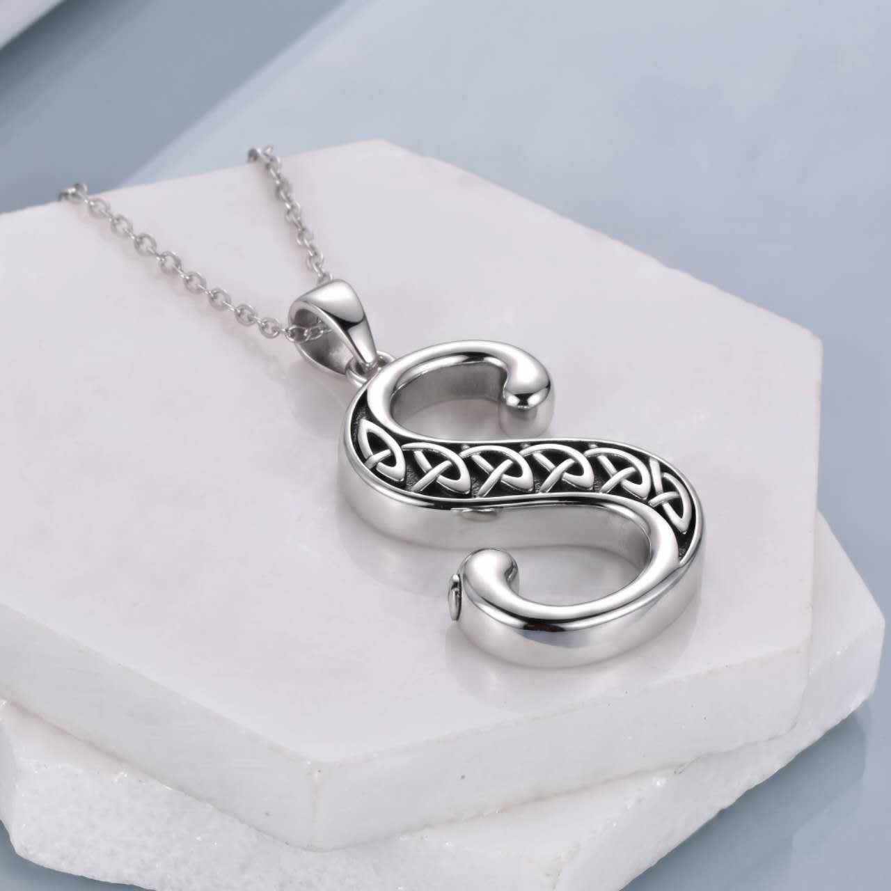 A silver pendant necklace shaped like the letter 'S', featuring an intricate Celtic knot design across the center, displayed on a white stone block.