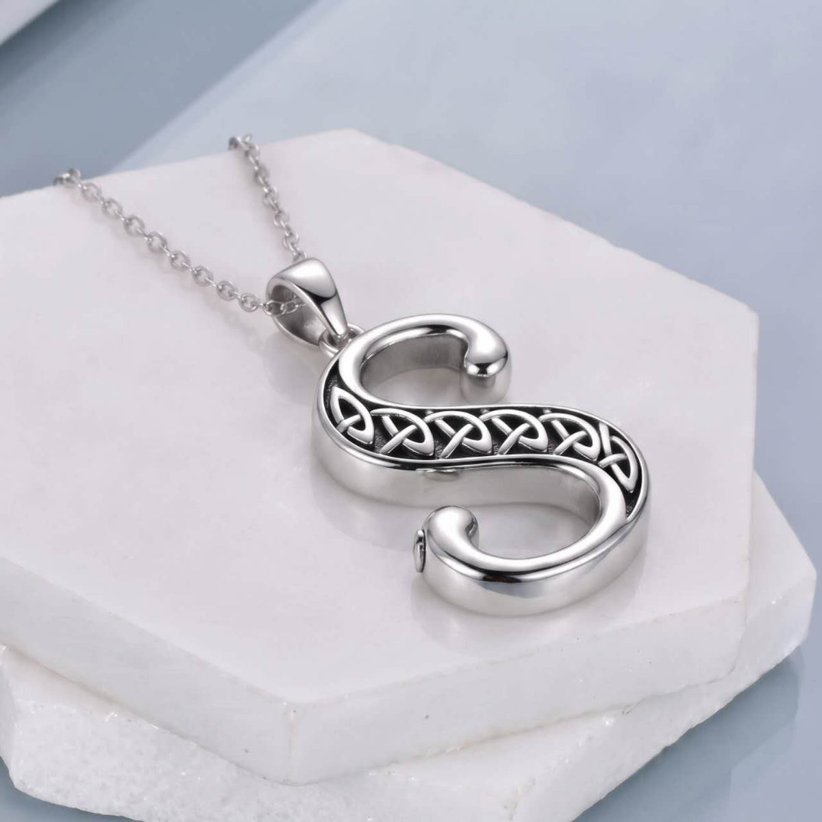 A silver pendant necklace shaped like the letter 'S', featuring an intricate Celtic knot design across the center, displayed on a white stone block.