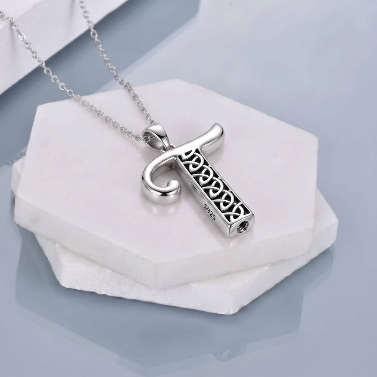 A silver-toned pendant necklace featuring a stylized letter 'T' with an openwork Celtic knot pattern inside, resting on stacked white hexagonal stone slabs against a soft light blue-gray background.