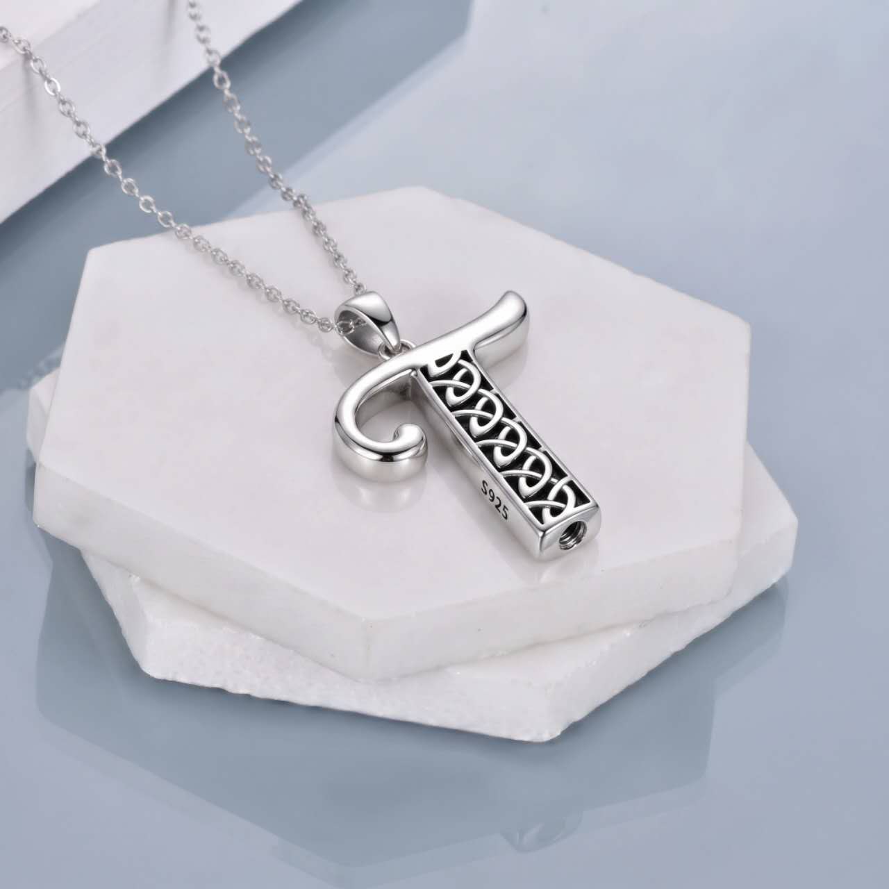 A silver-toned pendant necklace featuring a stylized letter 'T' with an openwork Celtic knot pattern inside, resting on stacked white hexagonal stone slabs against a soft light blue-gray background.