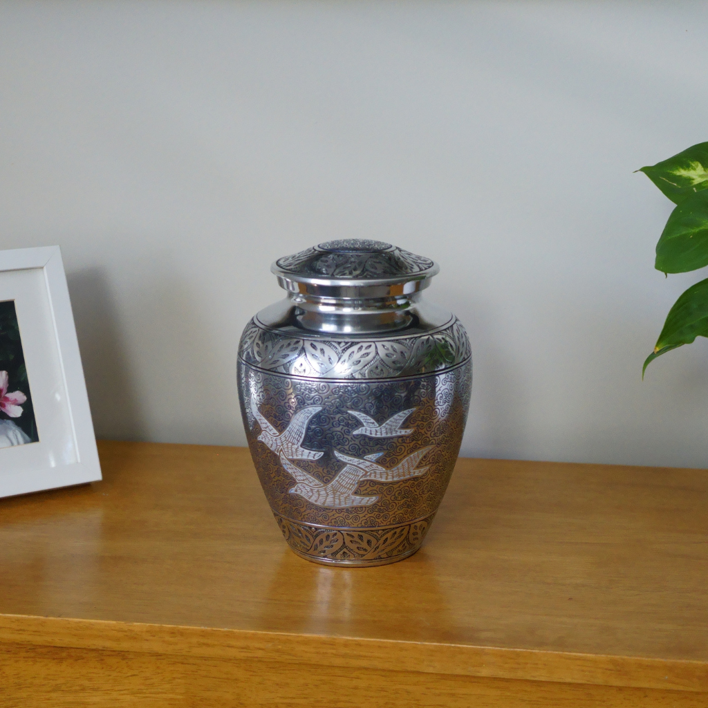 Silver urn with intricate cloud detailing and four prominent white birds etched in natural setting